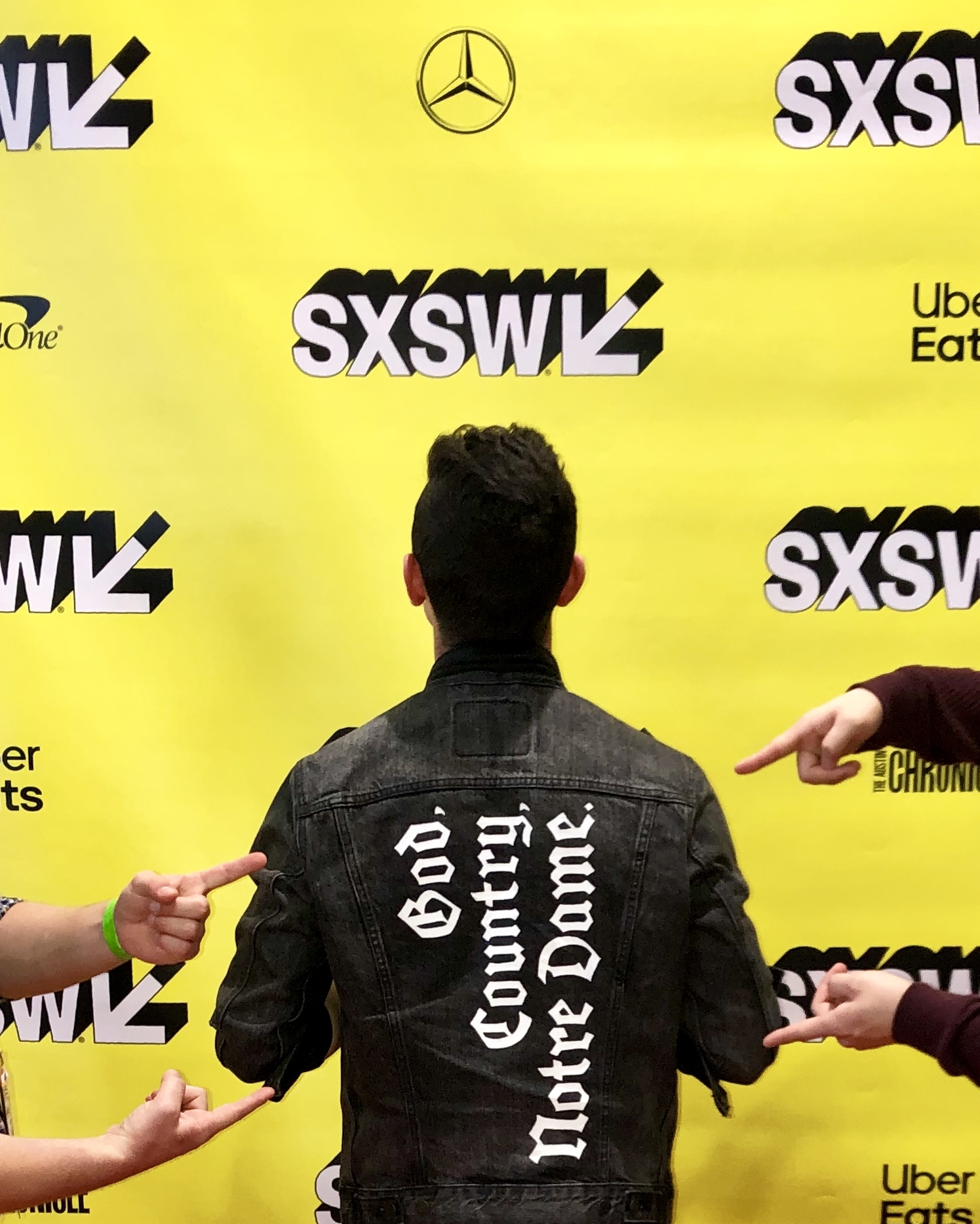 fingers point to the back of a jacket on a man reading: God. Country. Notre Dame. He stands in front of a SXSW backdrop