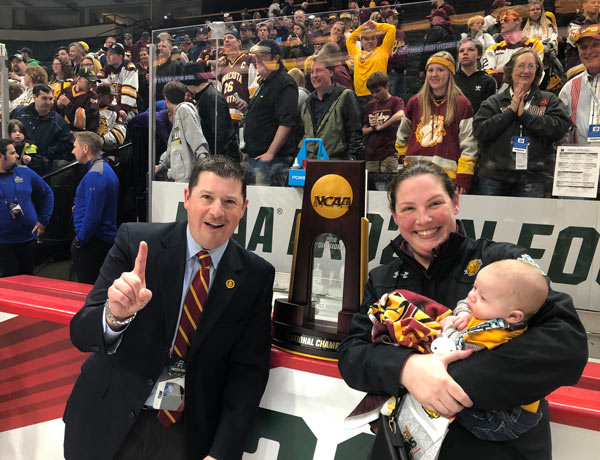 man and a woman holding a baby at a hockey game with an NCAA trophy