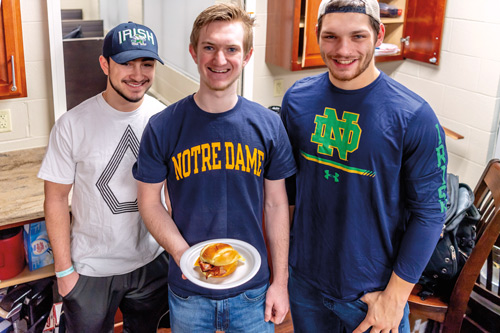three guys stand together, the one in the center holds a breakfast sandwich on a plate