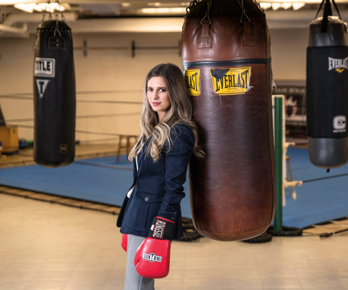 a woman in a business suit wears red boxing gloves and leans against a punching bag
