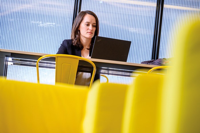 a woman sits at a table with a laptop in a room with yellow industrial furniture