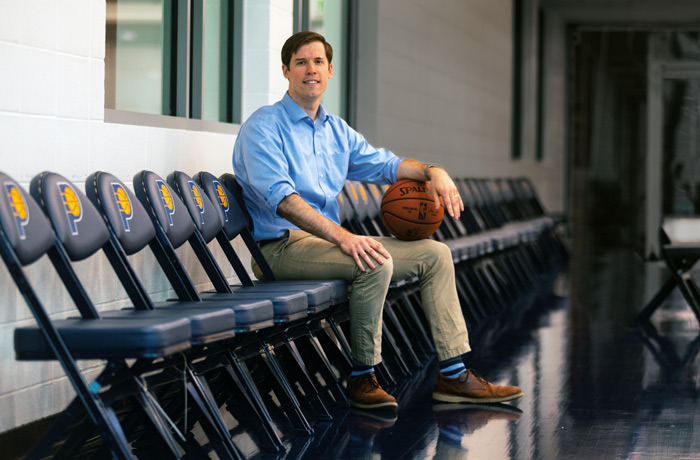 Peter Zanca sits on a row of chairs holding a basketball in the Indiana Pacers gym